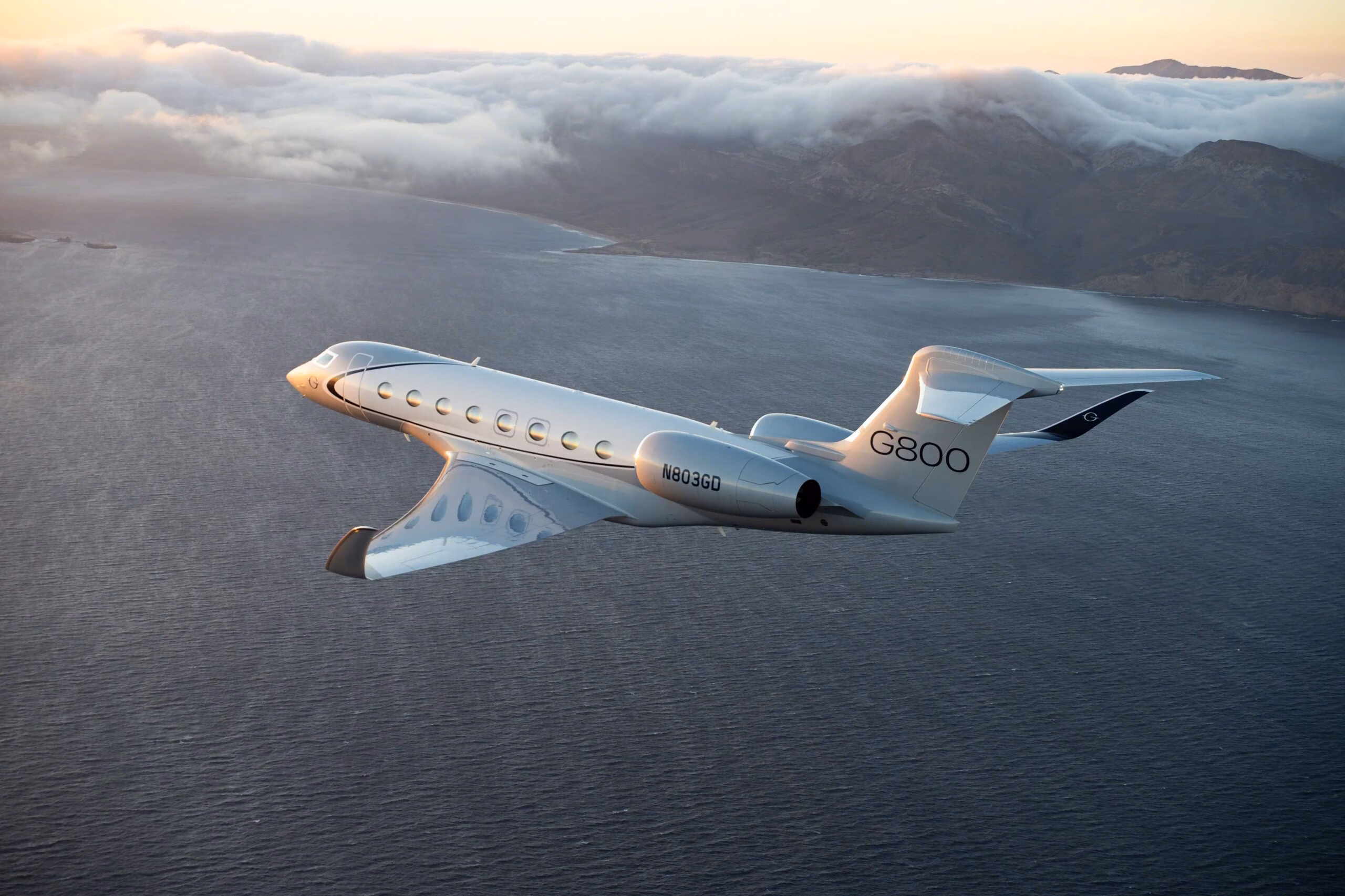 Gulfstream G800 flying over the ocean at sunrise with mountains and low clouds in the background.